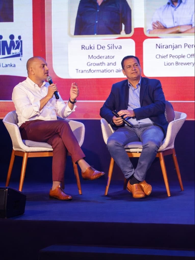 Two men sit on chairs during conference, holding microphones , with a conference backdrop behind them.